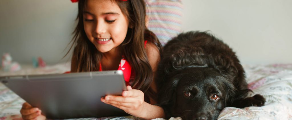 Little daughter in a red dress watching online videos while laying next to a black dog