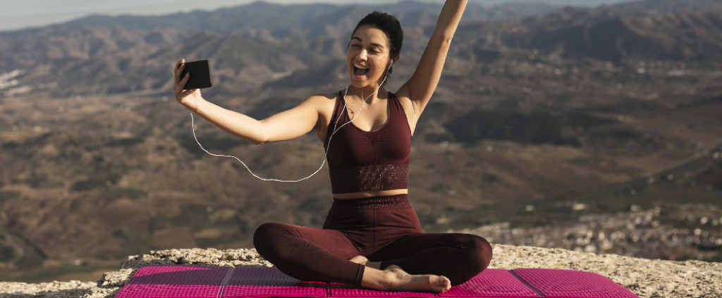 Woman in burgundy-colored training dress video calls on the top of a mountain.